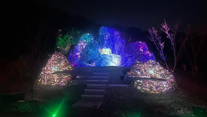 Large rock formations illuminated by colorful projections at night, creating a striking visual contrast against the dark sky.