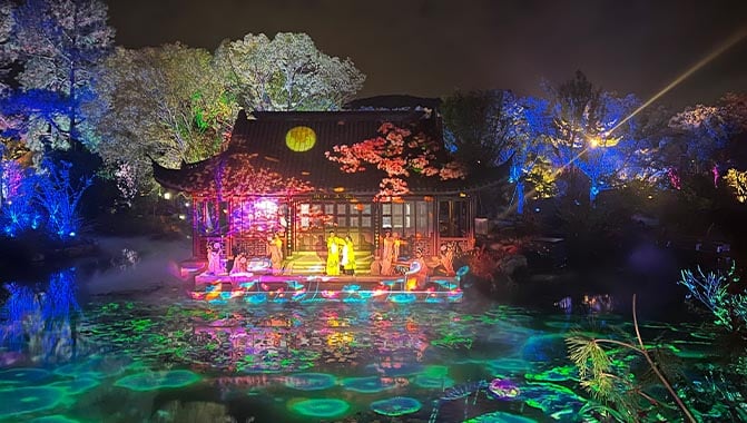 A picturesque pond with glowing lights, set against a backdrop of a building featuring projection displays and live performers, reflecting a calm atmosphere.