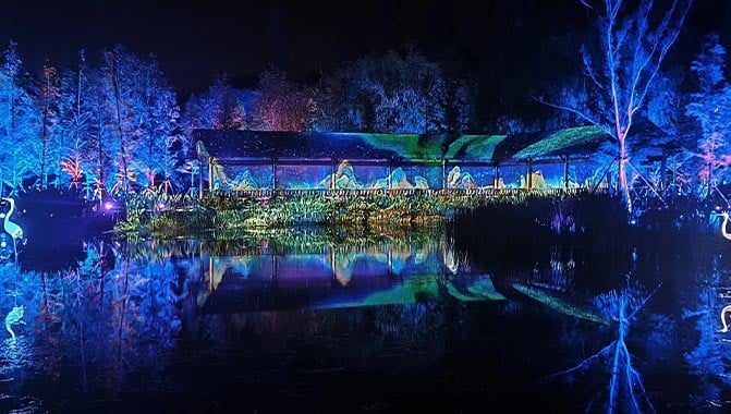 A tranquil lake with trees glowing in shades of blue and purple and projections on a thatched hut, creating a magical evening atmosphere.